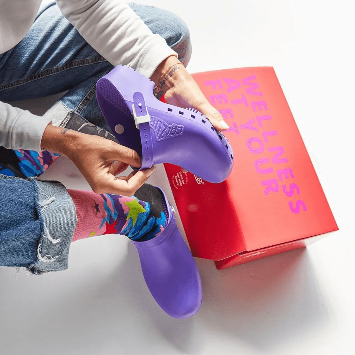 Purple clogs with adjustable strap, paired with colorful patterned socks, on denim jeans and a vibrant red box.
