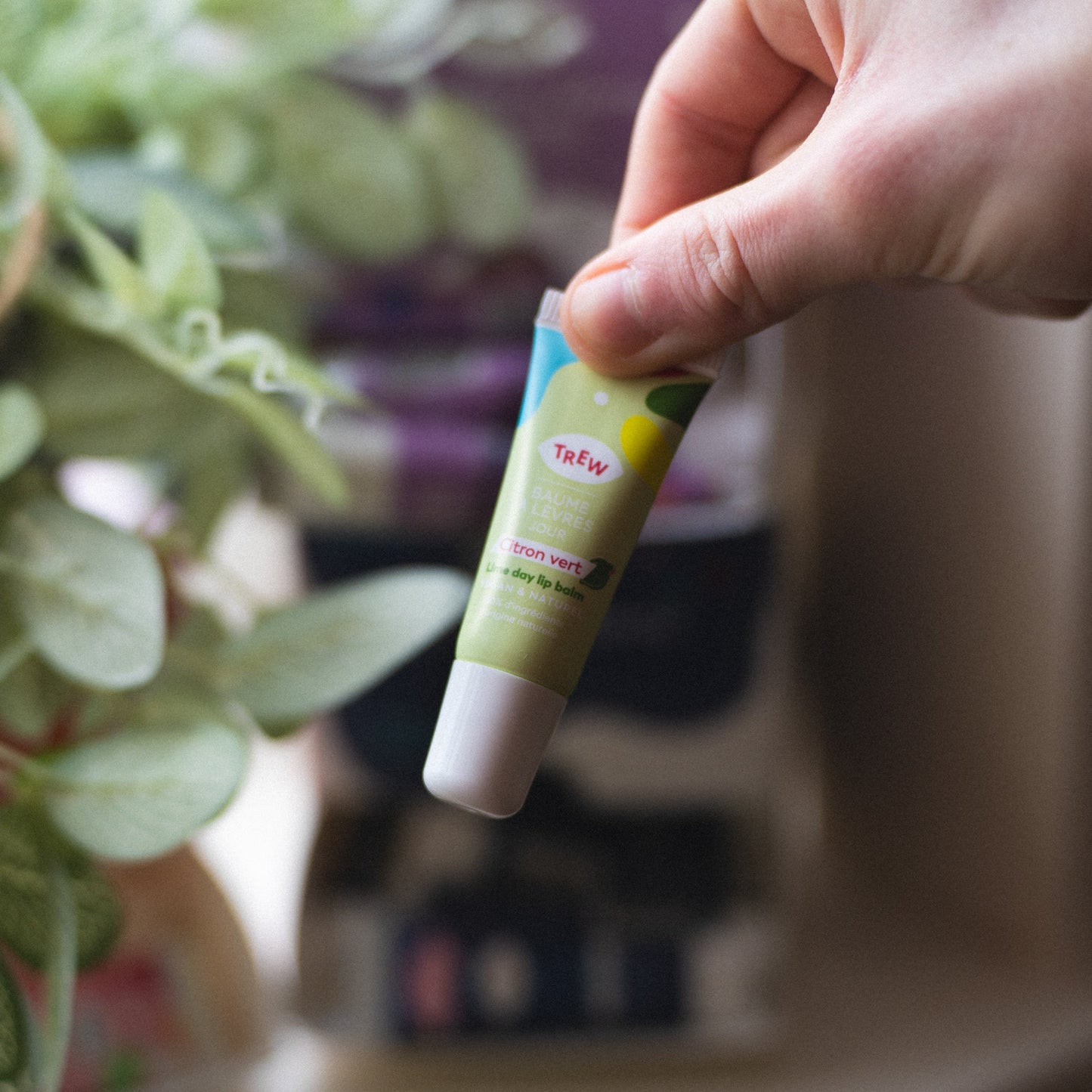 A hand holds a lime green tube of Trew Baume Jour Citron Vert lip balm against a blurred botanical background.