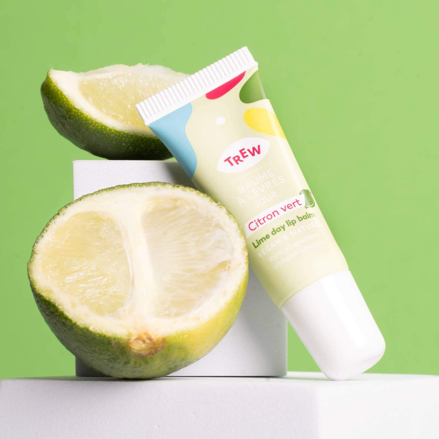 A tube of lime lip balm rests against white blocks with fresh lime halves on a bright green background.