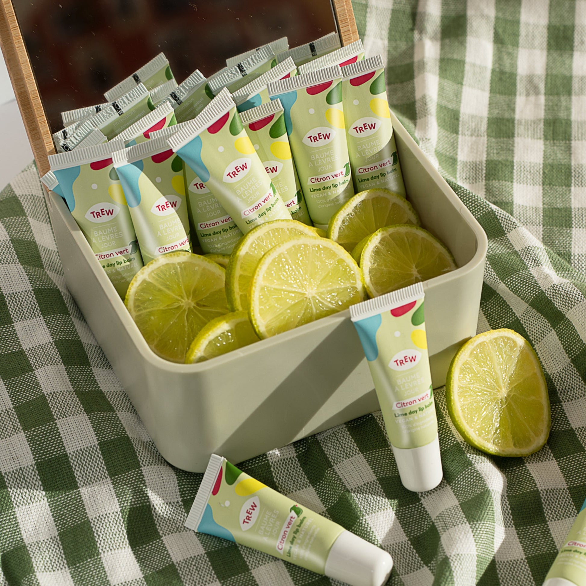 Lime lip balm tubes in a light green box with fresh lime slices, set on a green and white checkered fabric background.