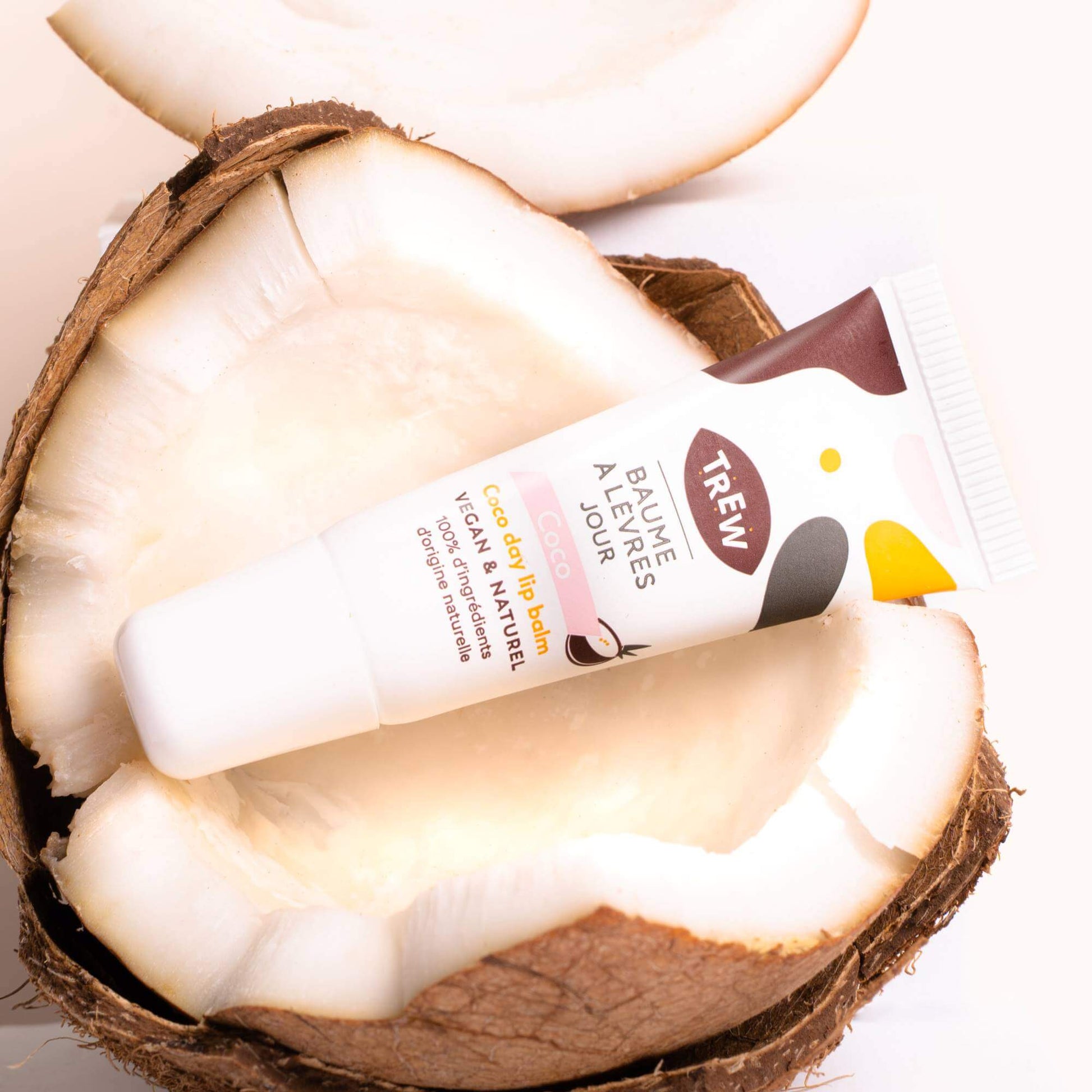 Tube de baume à lèvres Trew posé dans une noix de coco ouverte sur fond clair. Design blanc avec touches colorées.