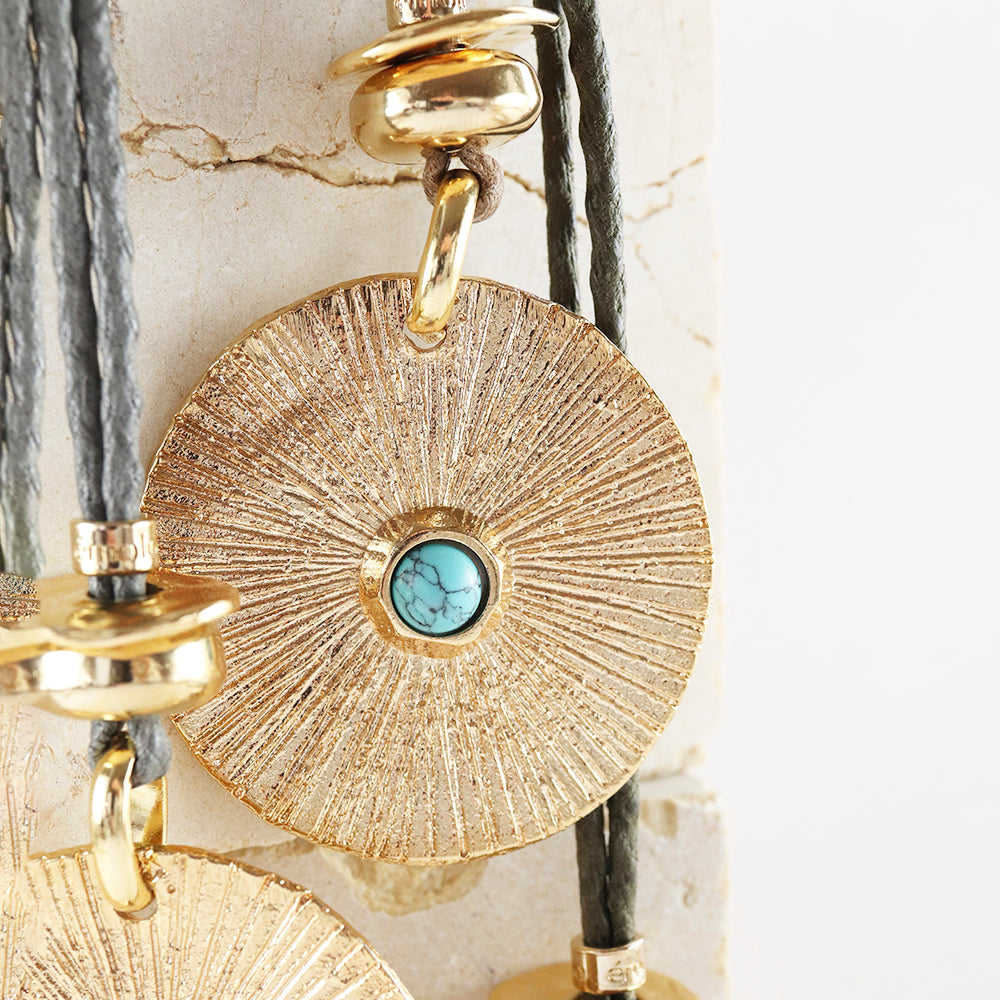 Collar de disco dorado con textura y piedra turquesa, cuerdas grises y cierre dorado. Estilo bohemio y elegante.