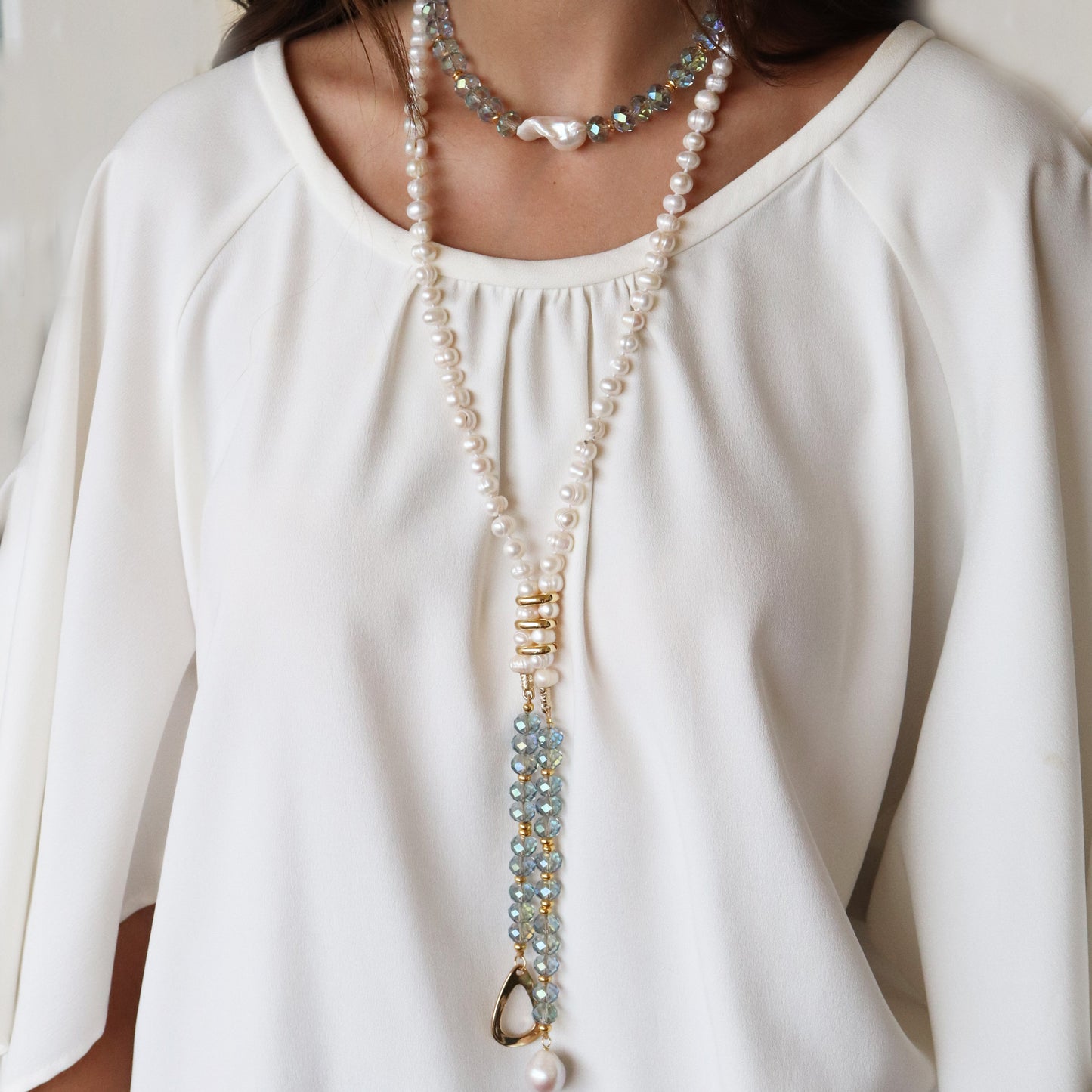 Collar largo de perlas blancas y piedras azules, con detalles dorados, sobre blusa blanca elegante.