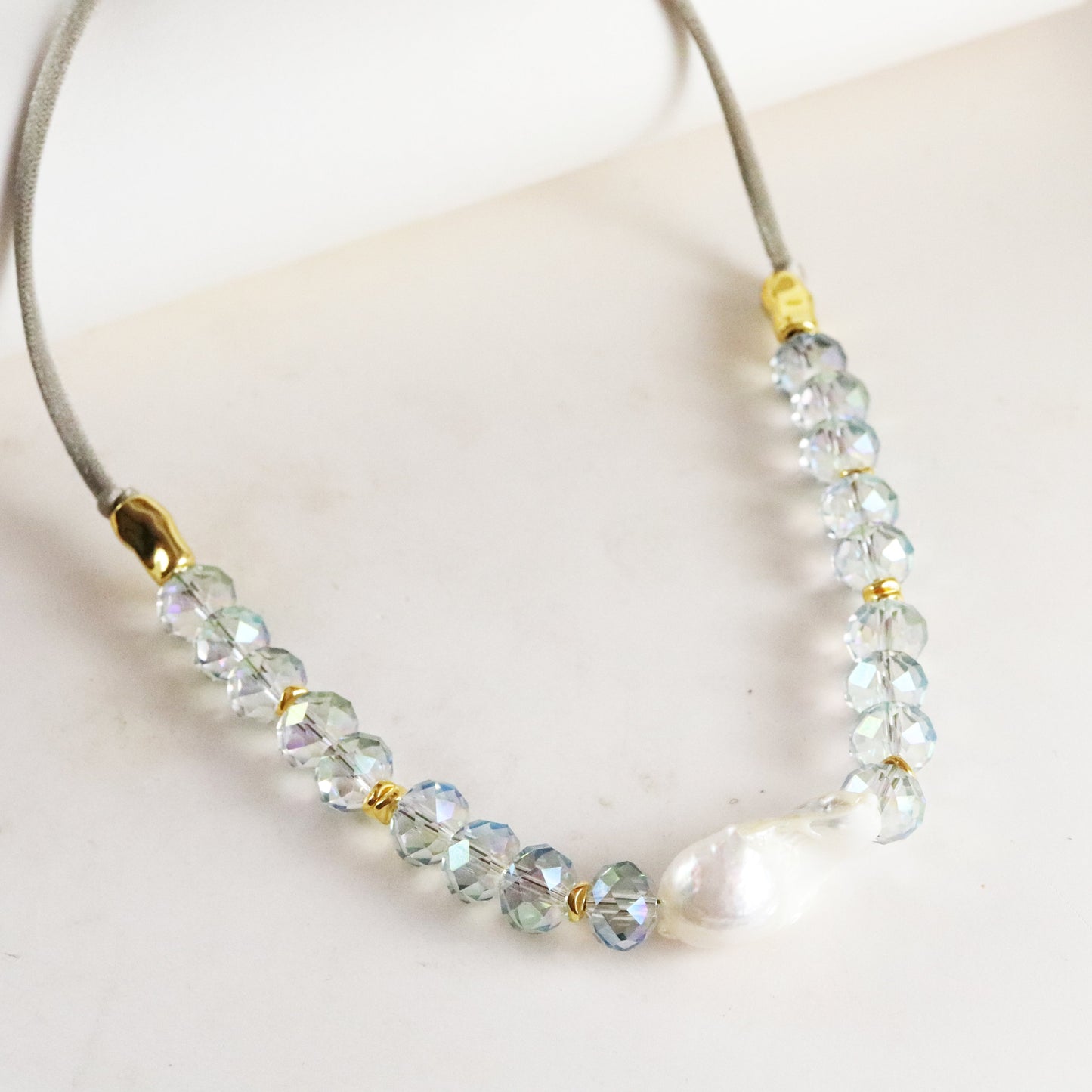 Collar elegante de cristal azul y perla blanca, con detalles en dorado y cordón gris claro.
