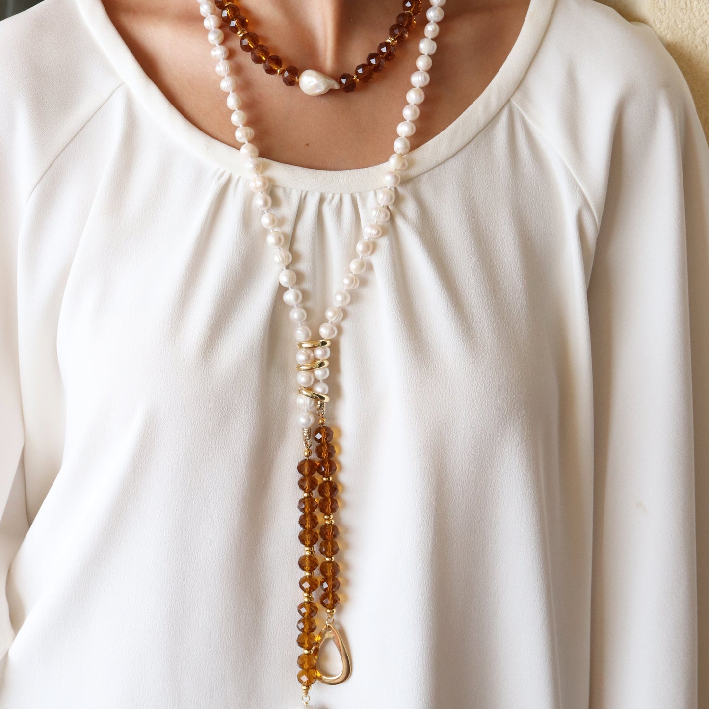 Collar largo de perlas y cristal ámbar, estilo bohemio, con detalles dorados y un diseño elegante sobre blusa blanca.