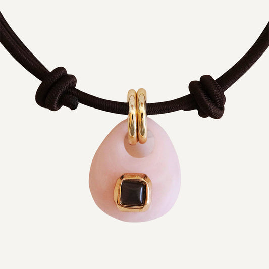 Collar de piedra rosa con detalle negro, adornado con aros dorados y cordón marrón, estilo elegante y moderno.