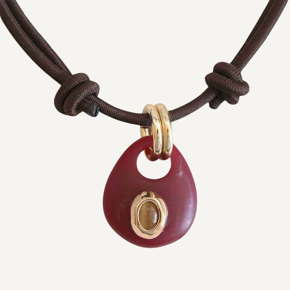 Colgante rojo en forma de gota con detalle dorado, cuerda marrón, estilo moderno y artesanal.