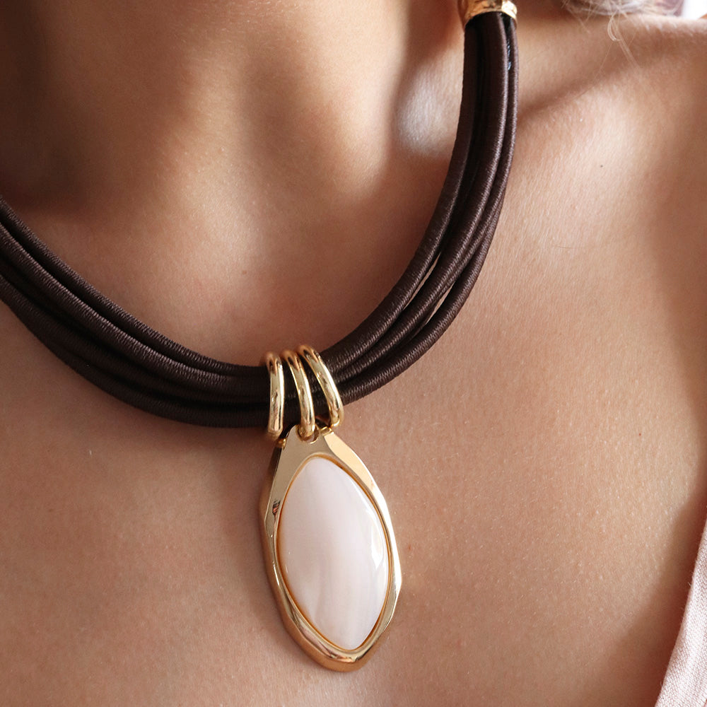Collar de acero dorado con piedra ovalada crema y cordón marrón, elegante y moderno.