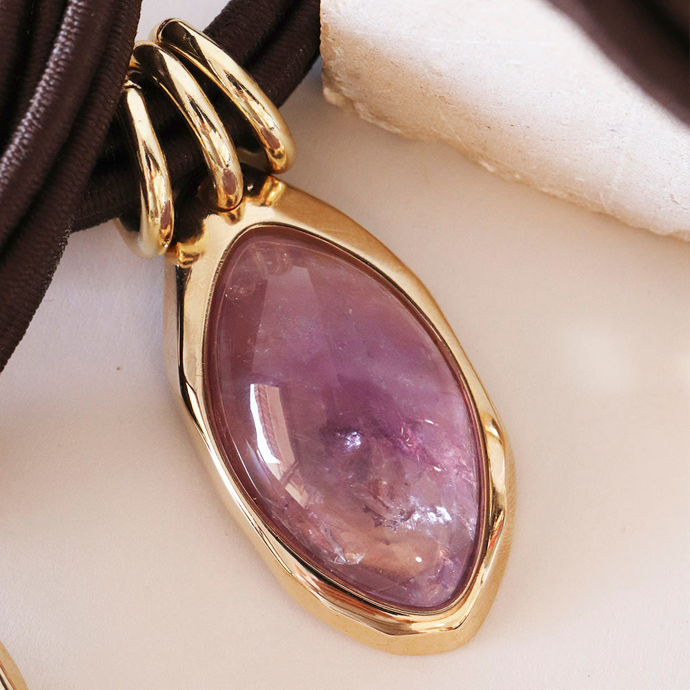 Colgante dorado con piedra amatista ovalada y cadena de cuero marrón. Ideal para estilos elegantes y bohemios.