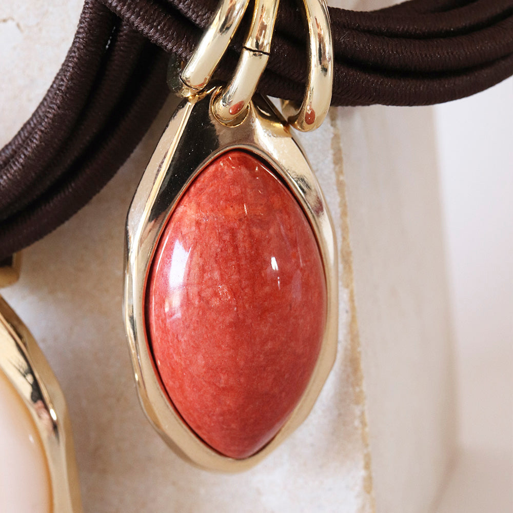 Collar con colgante ovalado de piedra roja y base dorada, sobre cuerda marrón, estilo moderno y elegante.