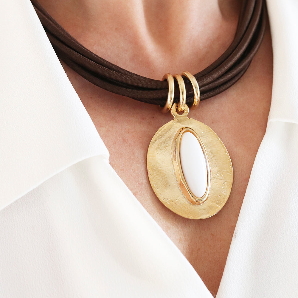 Collar de cuero marrón con colgante ovalado dorado y detalles en blanco, estilo moderno y elegante.