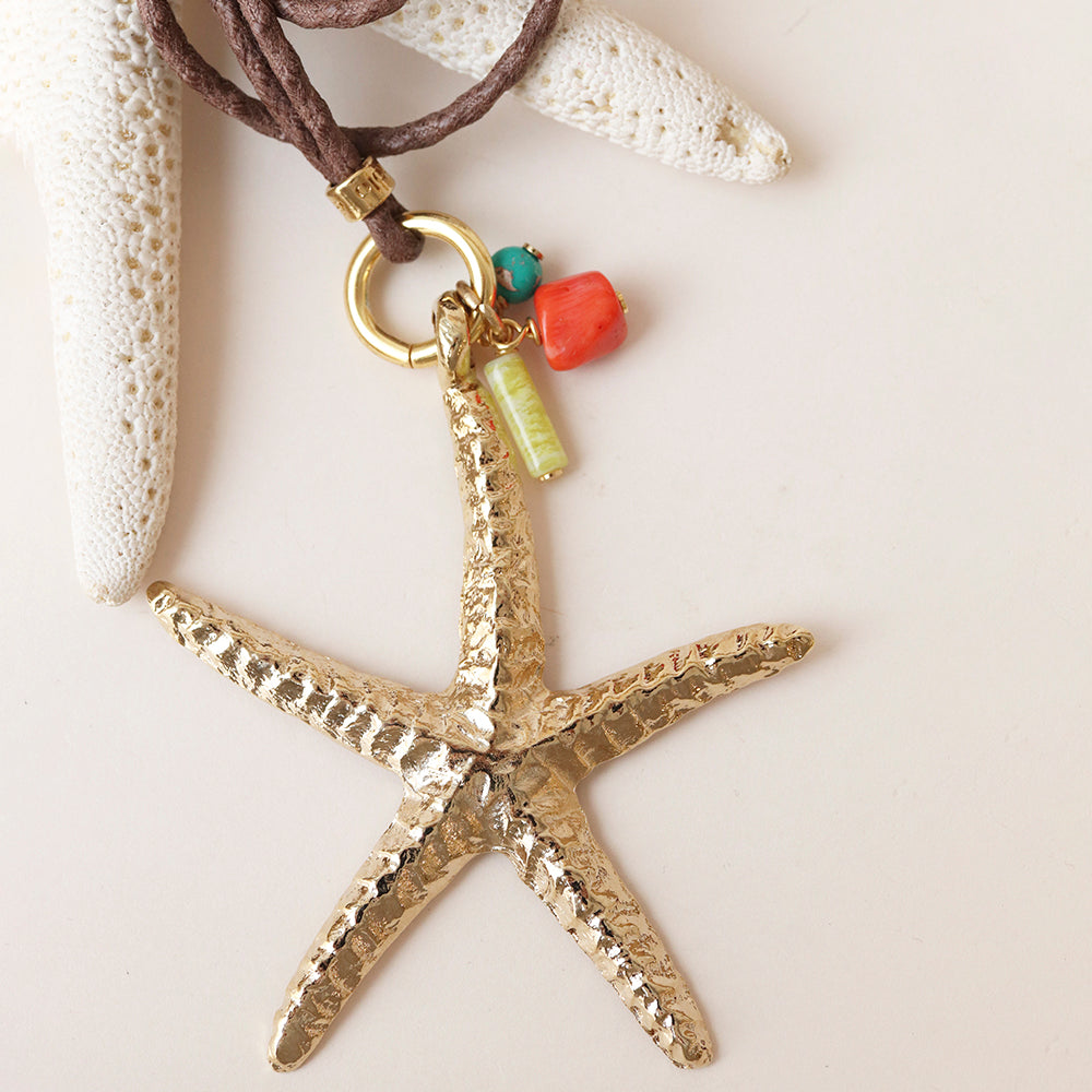 Collar con colgante de estrella de mar dorada, material metálico, con detalles en coral y turquesa sobre cordón marrón.