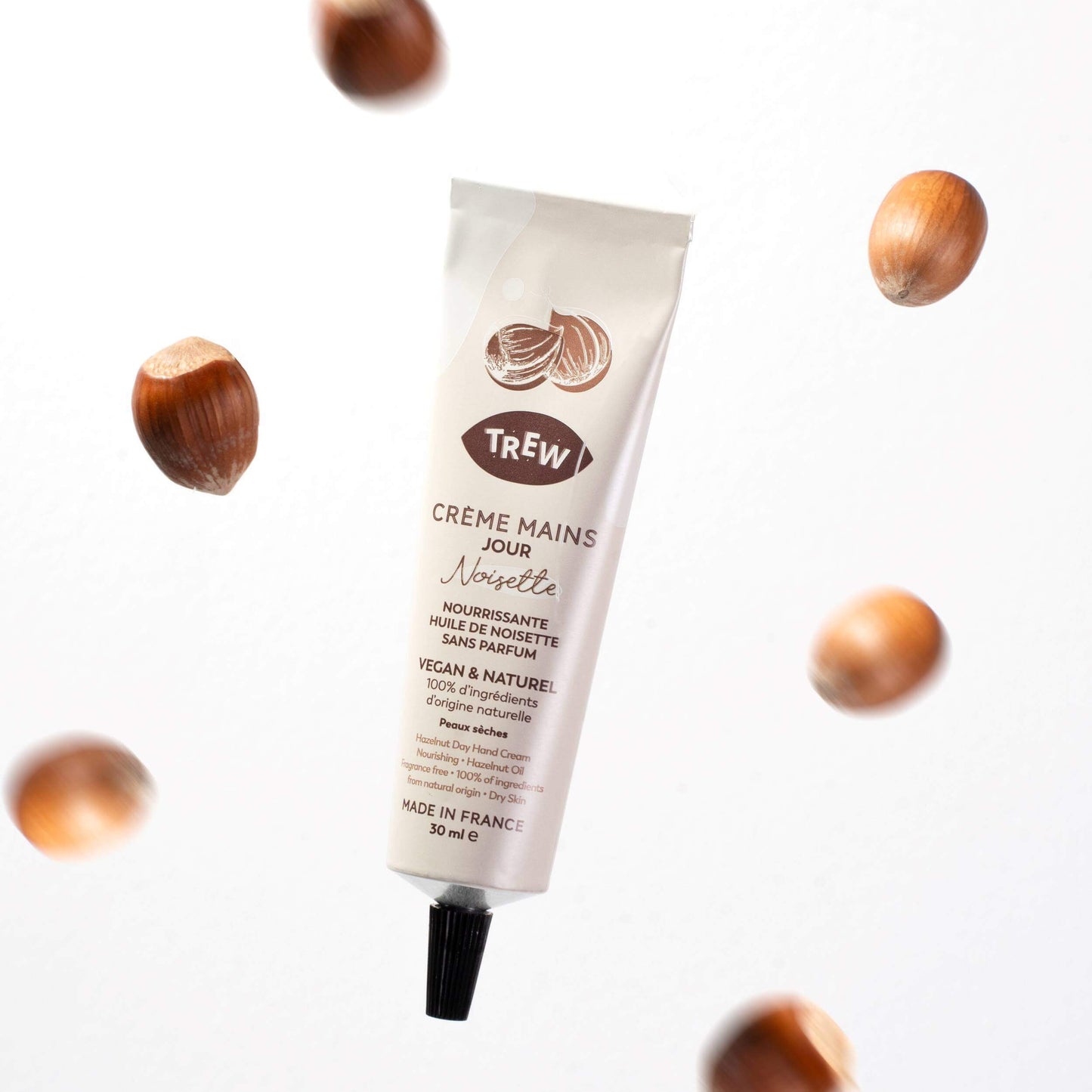 Beige tube of Trew hand cream surrounded by floating hazelnuts against a white background.