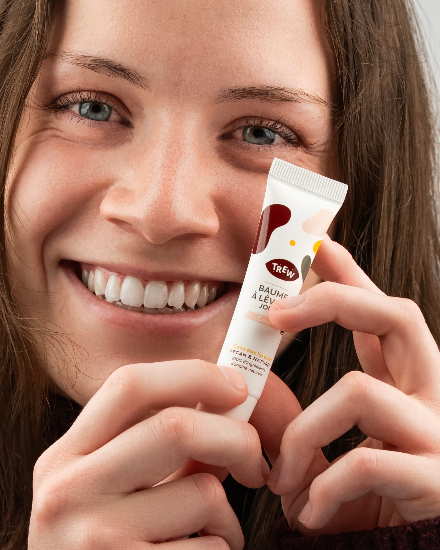 Jeune femme souriante tenant un tube de baume à lèvres vegan Trew Cosmetic à motifs colorés sur fond neutre.