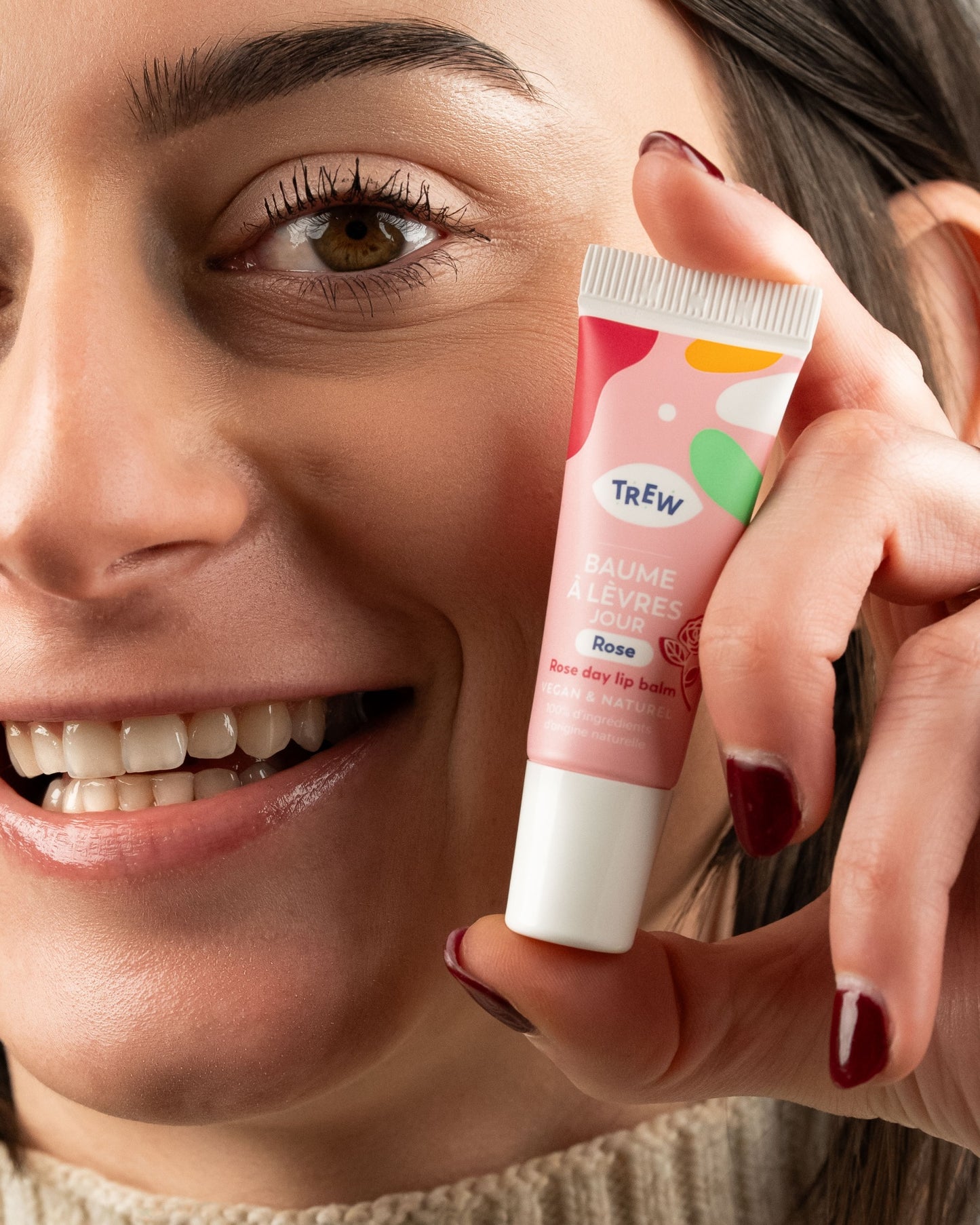 Smiling person holding a pink squeeze tube of Trew rose lip balm featuring abstract colorful shapes.