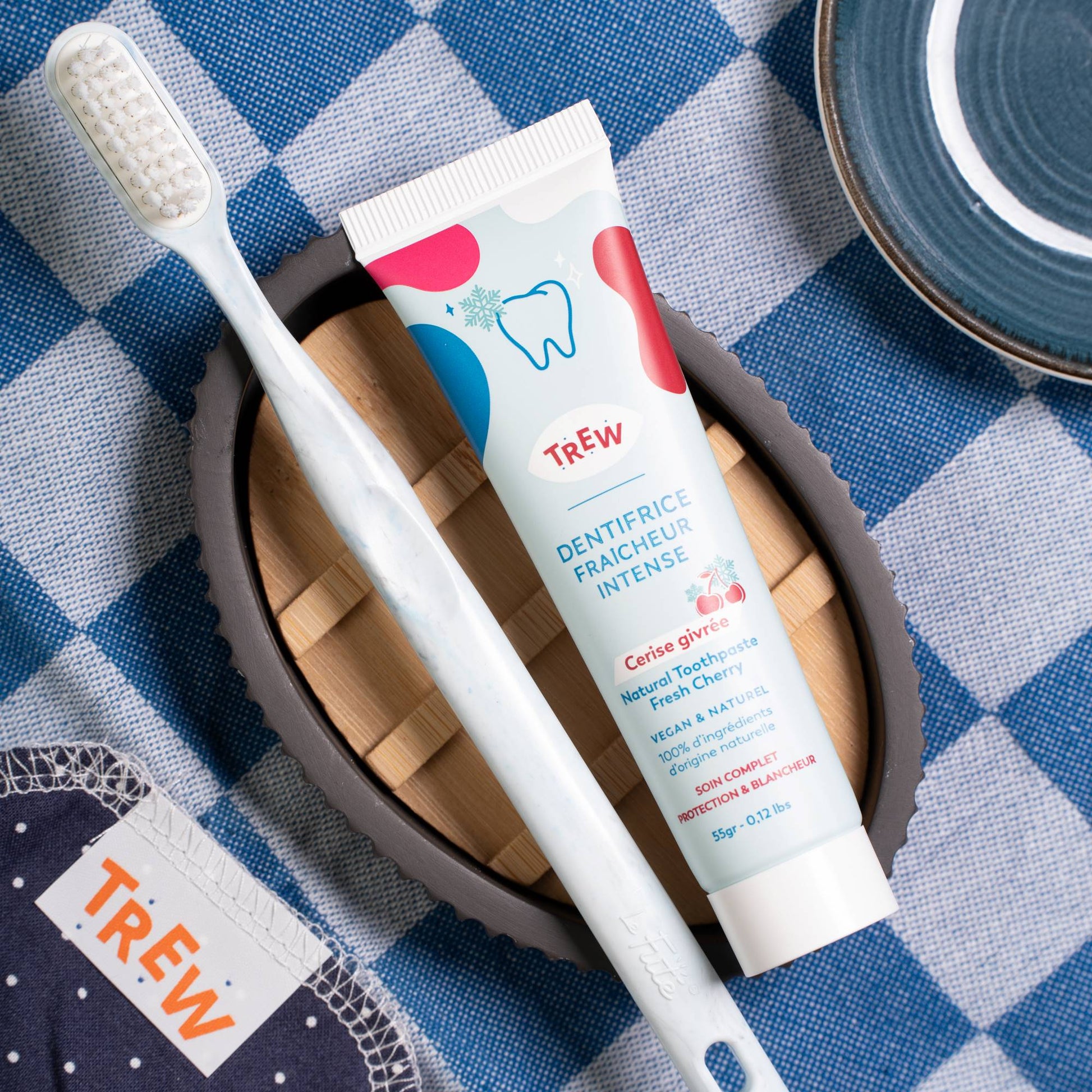 Tube de dentifrice Trew à la cerise et brosse à dents blanche sur un support en bois, posés sur une nappe à carreaux.