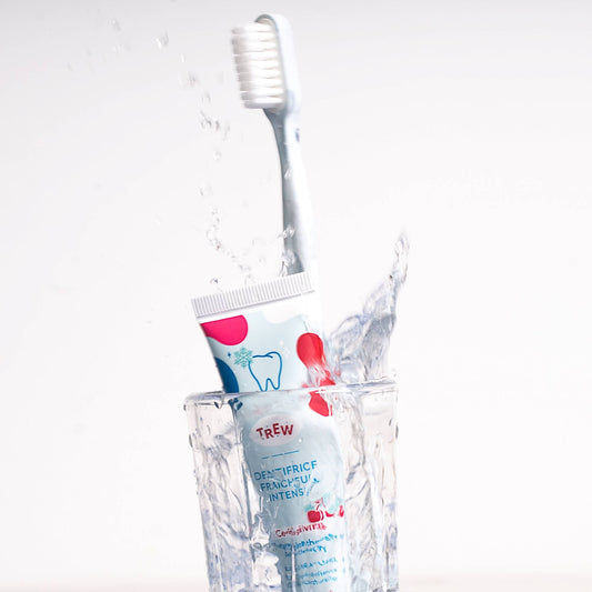 Tube de dentifrice Trew Cerise Givrée et brosse à dents dans un verre d'eau avec éclaboussures sur fond blanc.