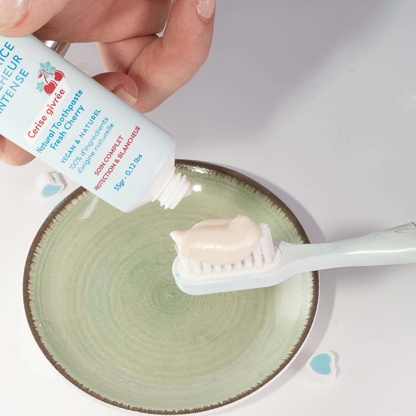 Main tenant un tube bleu pastel, on dépose une noisette de dentifrice beige crémeux sur une brosse à dents au-dessus.