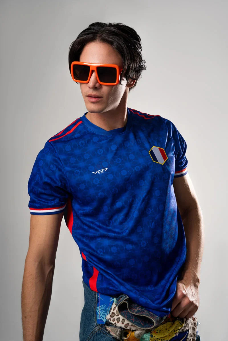 Maillot bleu VO7 à motif géométrique, col rond, détails tricolores et logo blanc sur la poitrine.