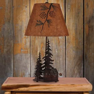 Rustic table lamp with a dark metal base featuring a cabin and pine trees, topped with a textured brown pinecone shade.