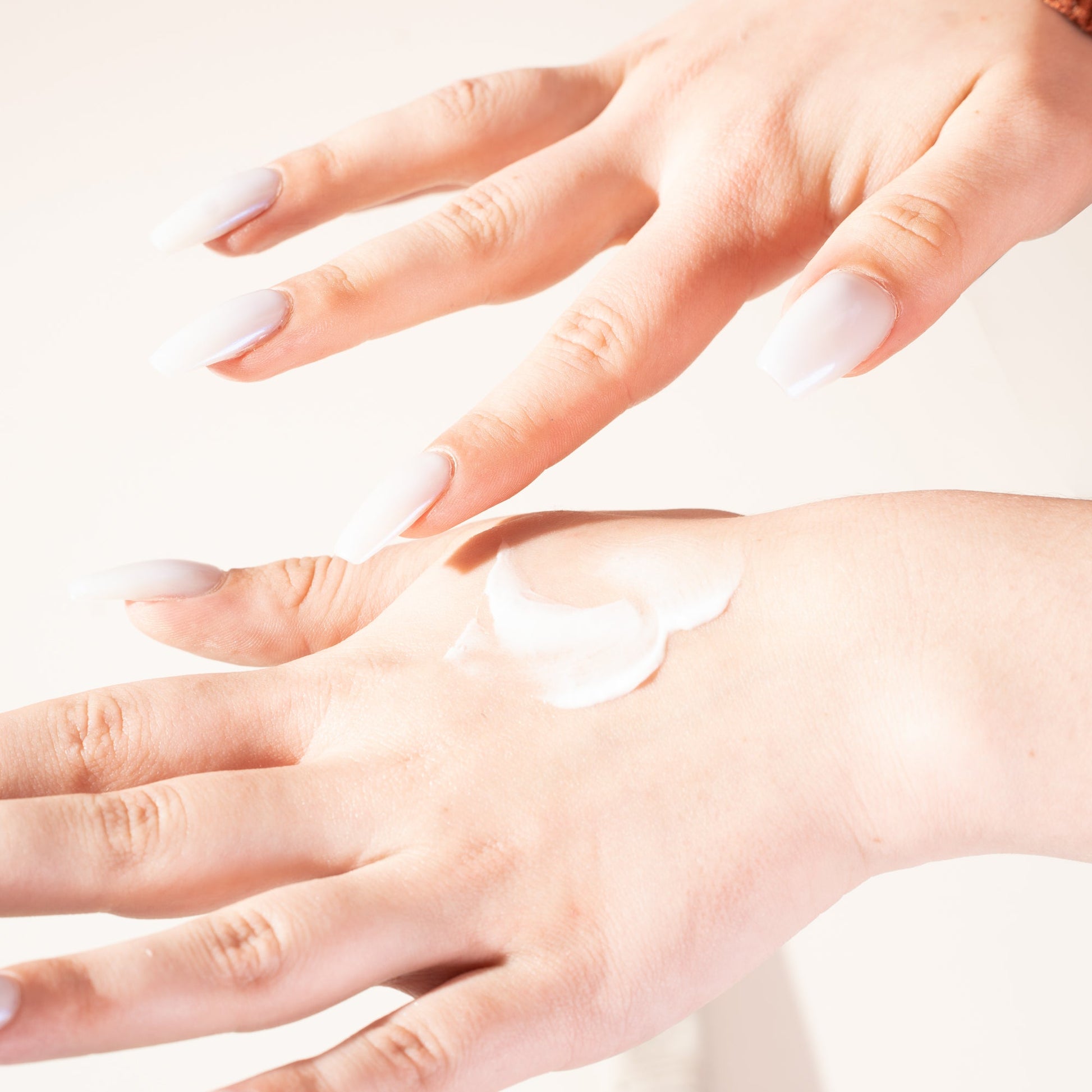 Creamy white hand cream dabbed on skin, hands with long glossy white nails against a soft neutral background.