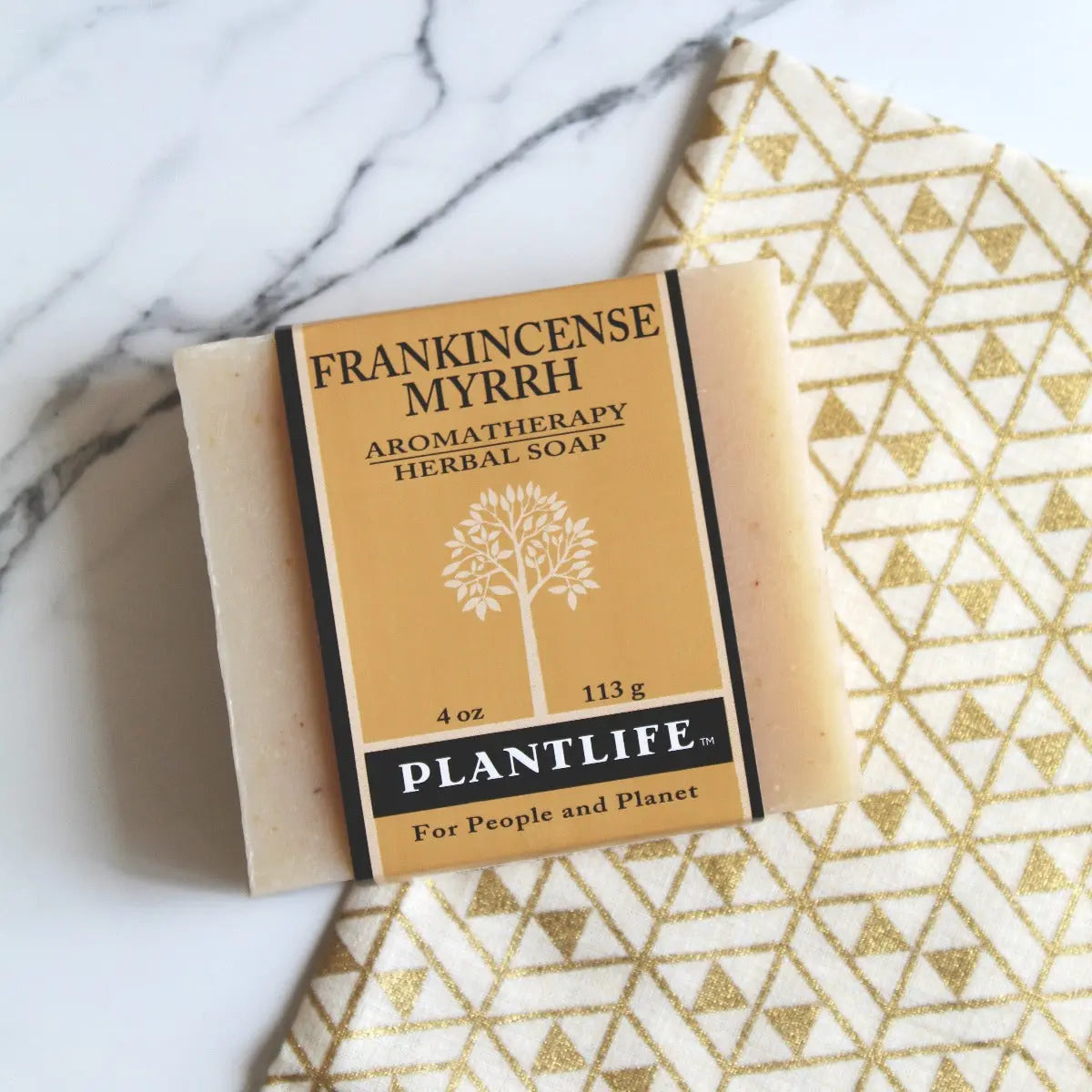 Frankincense Myrrh bar soap with a tan label featuring a white tree graphic, on a marble and gold geometric cloth.