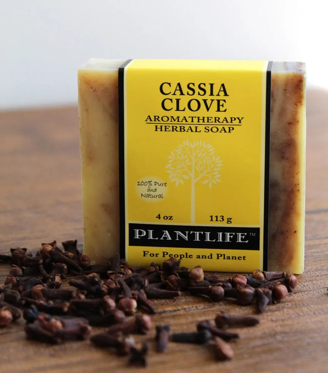 A bar of Cassia Clove soap in yellow packaging sits on a wooden surface beside a pile of dried whole cloves.