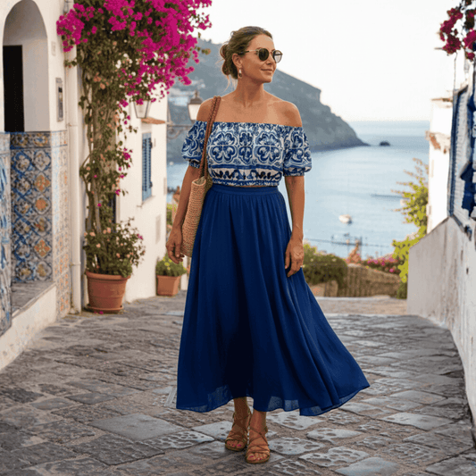 Woman wearing a blue and white Majolica print off-shoulder cotton top and a long flowy blue skirt on a coastal street.