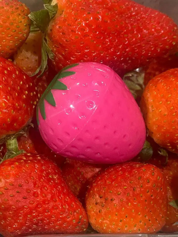 Bright pink, strawberry-shaped lip butter container nestled among a cluster of fresh, ripe red strawberries.
