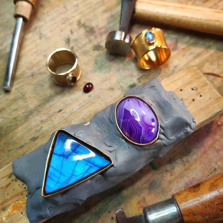 Anillo dorado con piedra azul triangular y piedra morada, sobre superficie de arcilla en taller de joyería.