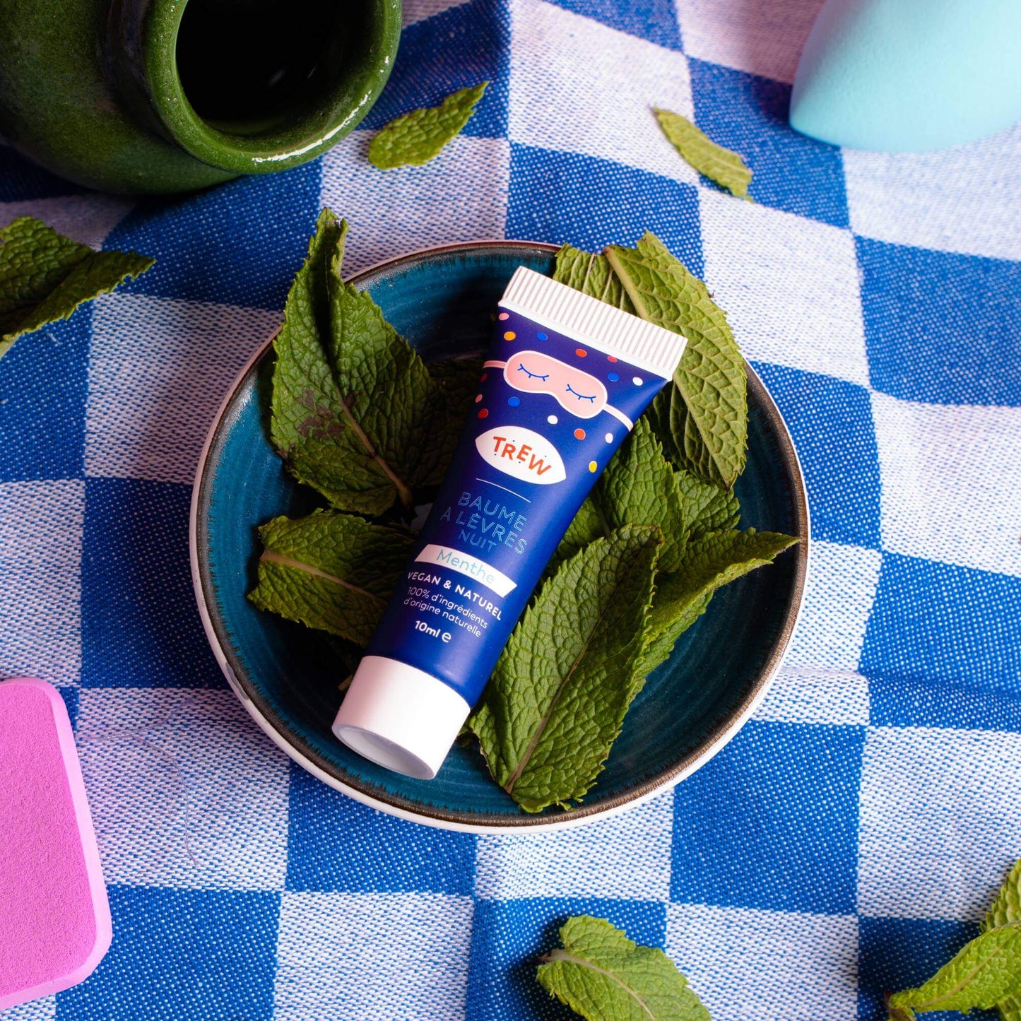 Tube de masque lèvres Trew bleu, posé sur des feuilles de menthe dans un bol, sur une nappe à carreaux bleus et blancs.