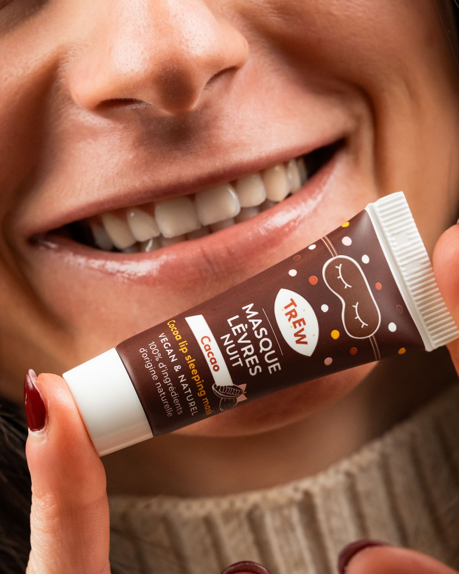 A person holding a brown tube of Cacao lip sleeping mask with a sleep mask icon, set against a smiling face.