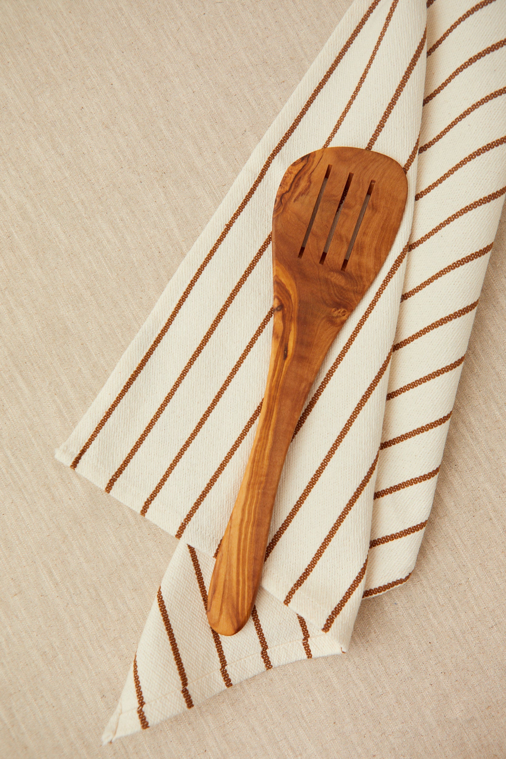 Espátula de madera de olivo, color marrón, sobre servilleta de algodón con rayas beige y marrón.