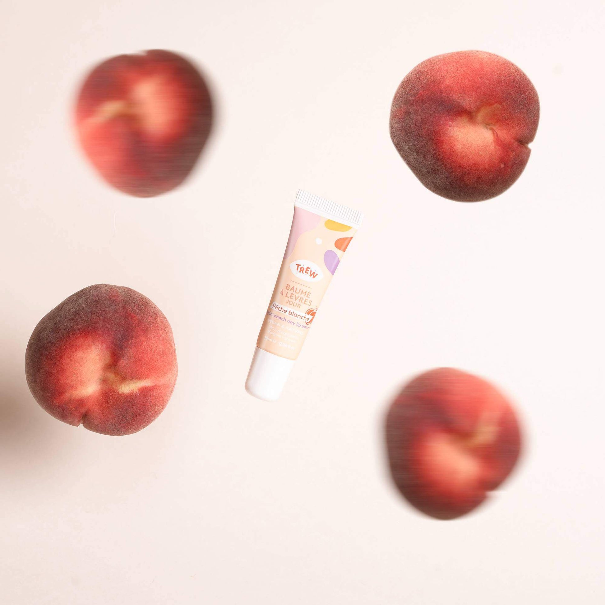 Trew "Pêche blanche" lip balm tube surrounded by floating fresh peaches against a soft pink background.