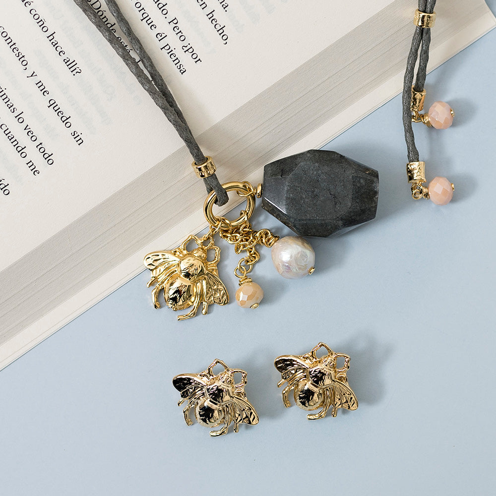 Collar gris con colgante negro, cuentas de perla y aretes dorados con diseño de abeja.