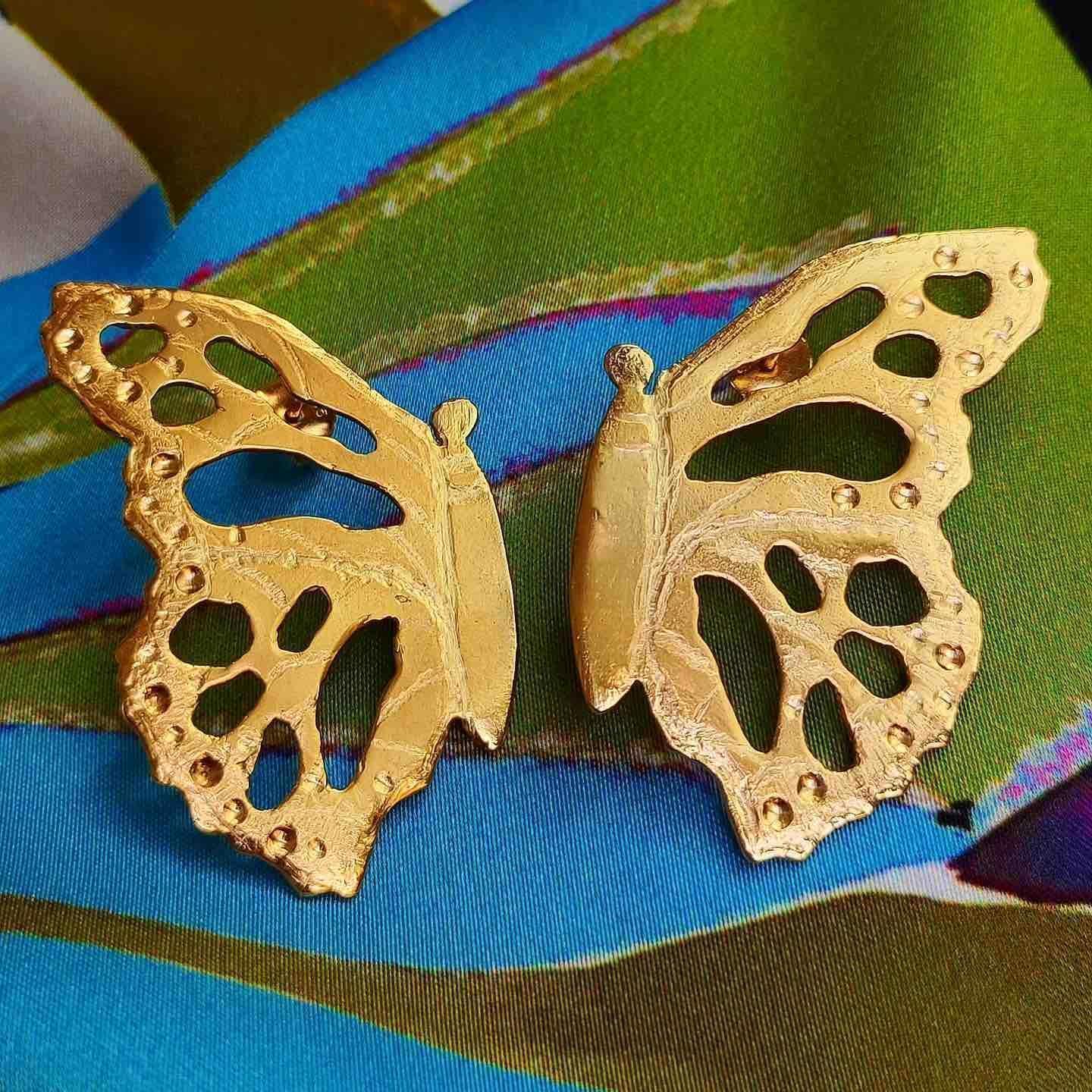 Broche de mariposa dorado de metal con diseño detallado, sobre fondo de seda colorido y vibrante.