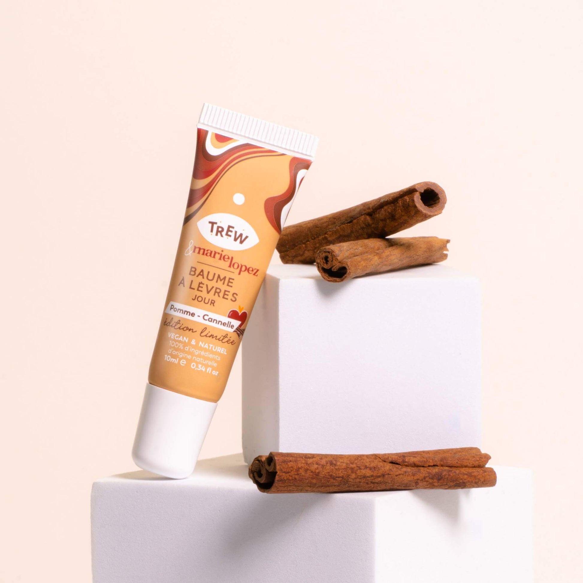 A tube of Trew x Marie Lopez Pomme Cannelle lip balm on a white cube with cinnamon sticks against a beige background.
