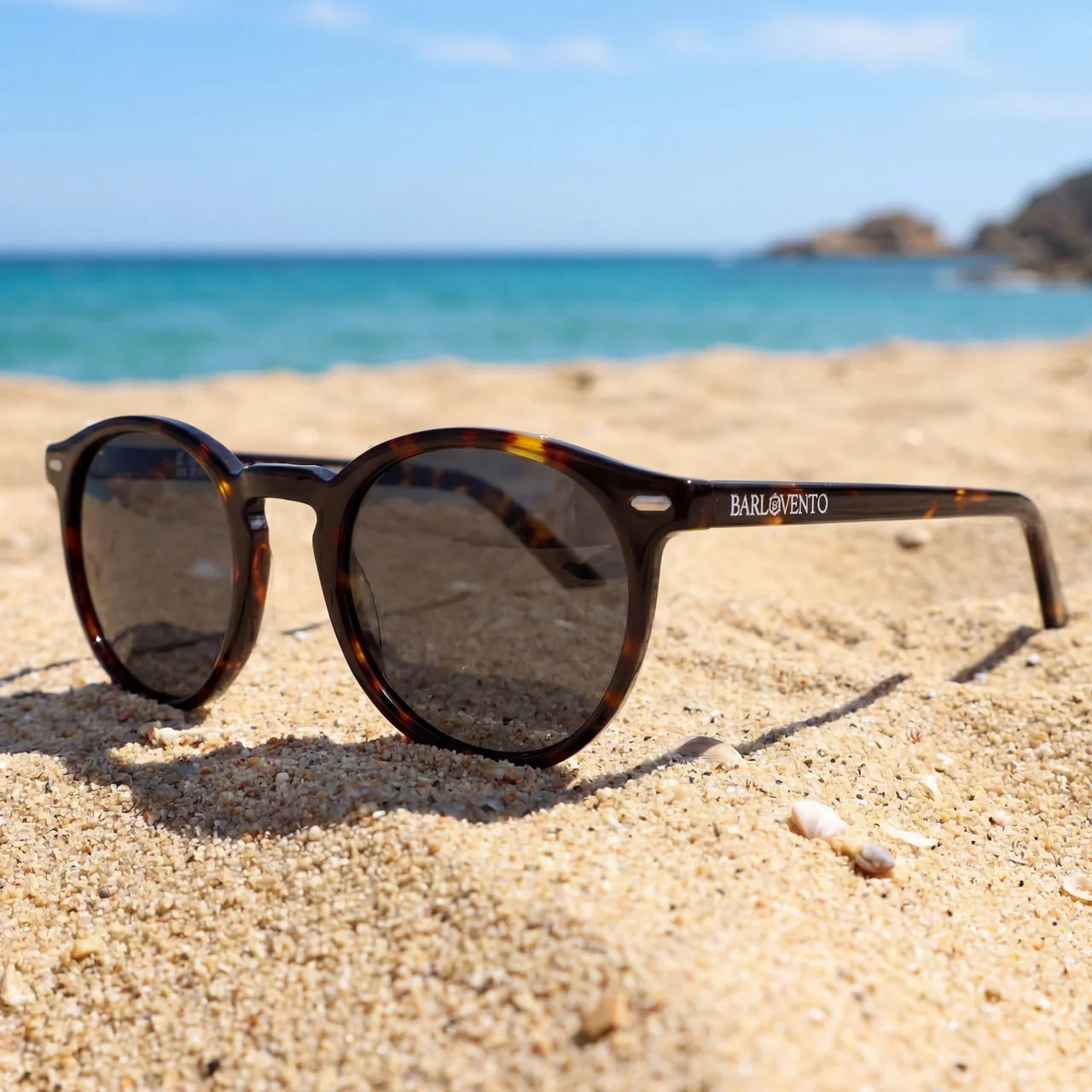 Gafas de sol redondas de carey con lentes polarizadas color marrón sobre la arena de la playa con el mar de fondo.