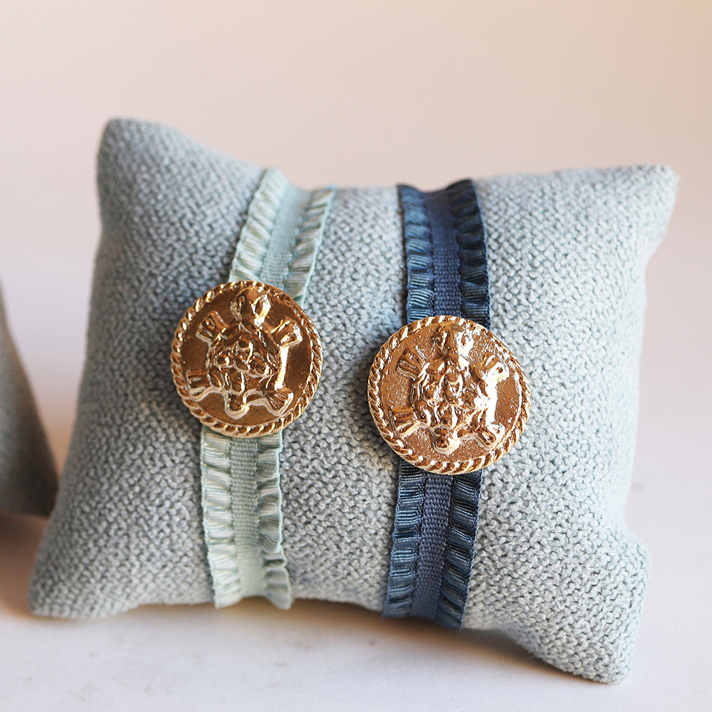 Conjunto de broches dorados en forma de moneda sobre cojín de tela azul y cintas de seda verde y azul.