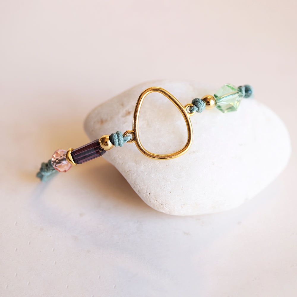 Pulsera de hilo azul, cuentas de cristal morado y verde, diseño moderno con detalle dorado en forma de gota.
