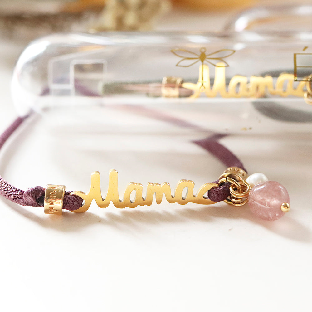 Collar dorado "Mama" con cinta morada y piedras rosa claro, presentación elegante en estuche plástico.