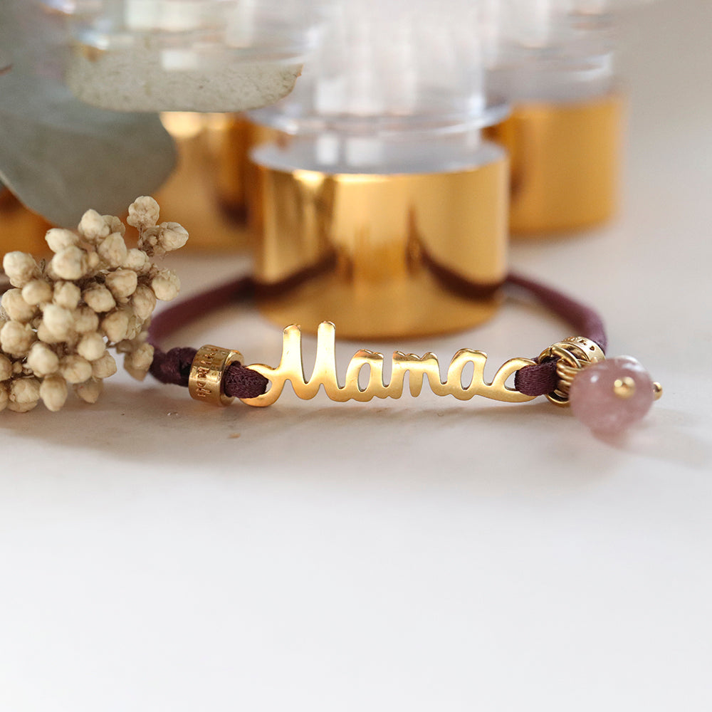 Pulsera dorada con el nombre "Mama" y charm de piedra rosa, sobre hilo marrón. Ideal como regalo.