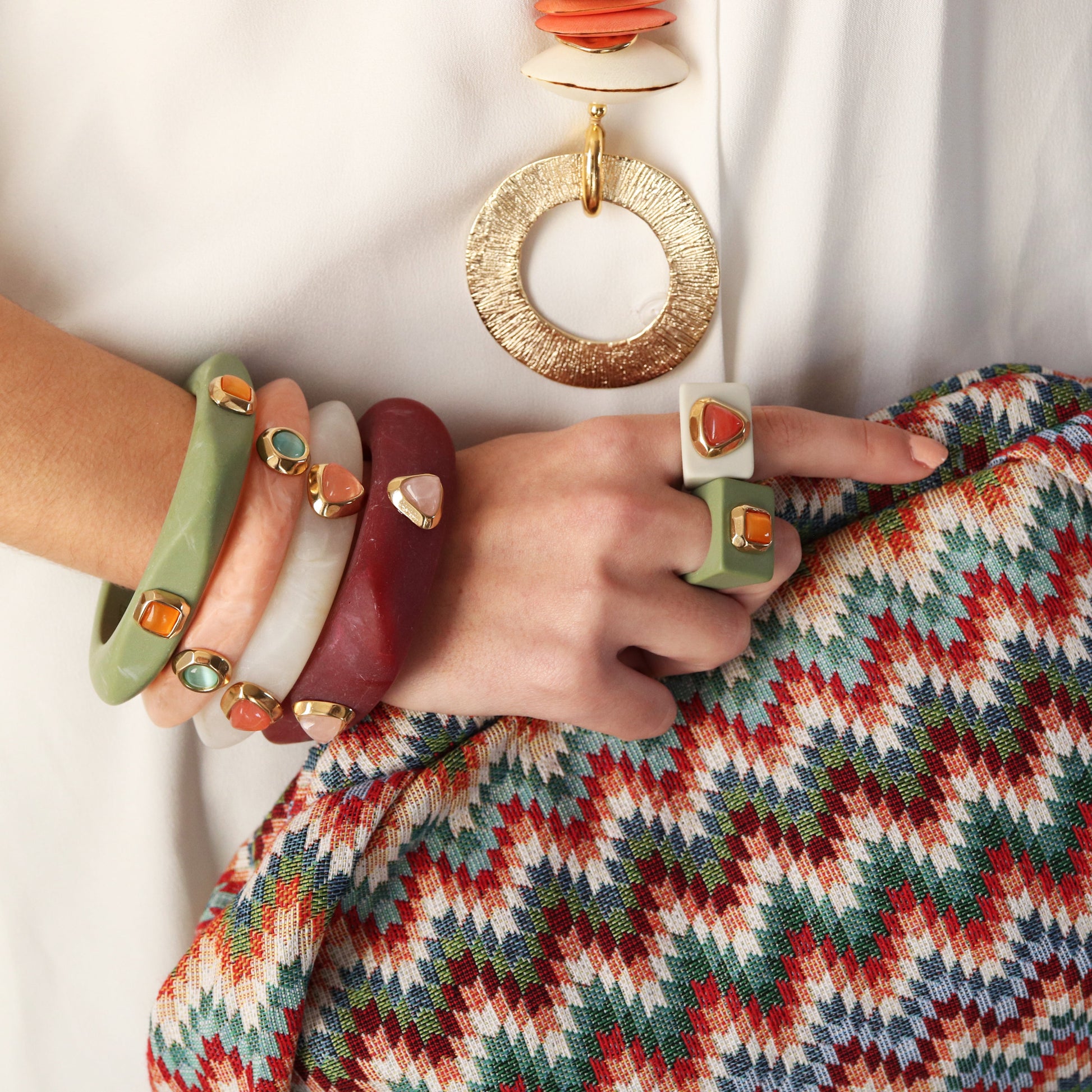 Brazaletes de plástico verde, rojo y beige con detalles dorados; anillo blanco con gemas. Estilo bohemio y vibrante.
