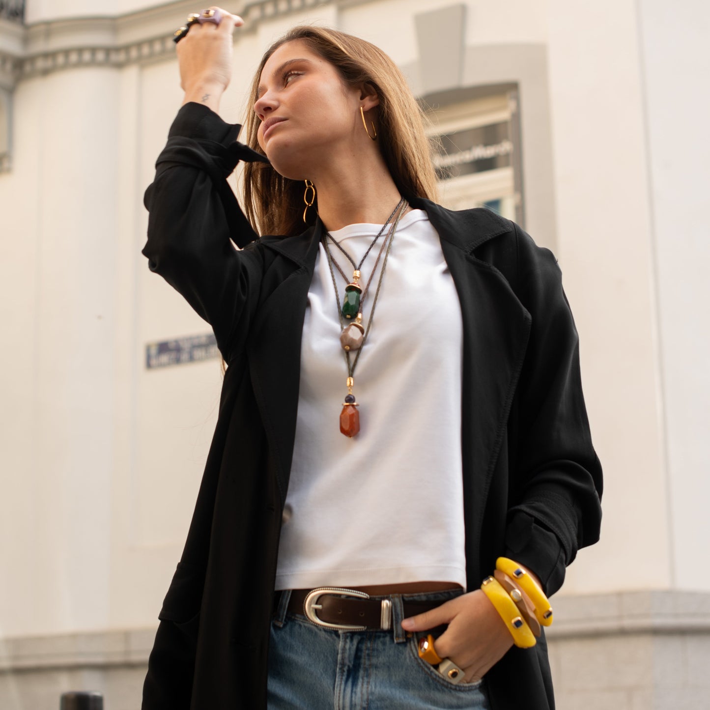 Mujer con abrigo negro, blusa blanca y jeans; accesorios de piedras y pulseras amarillas. Estilo urbano y elegante.