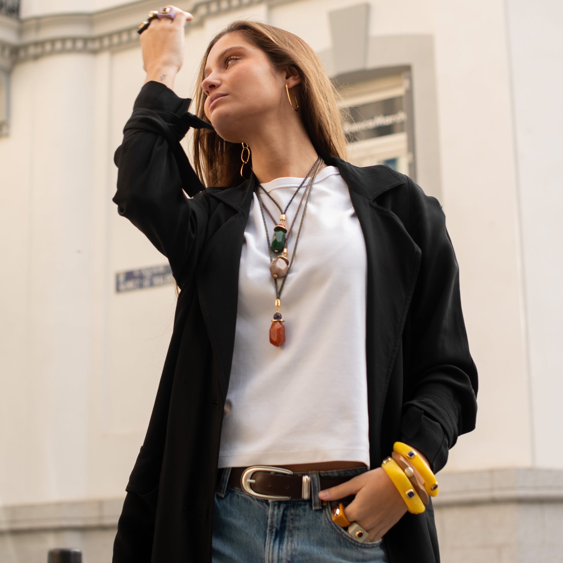 Mujer con abrigo negro, blusa blanca y jeans; accesorios de piedras y pulseras amarillas. Estilo urbano y elegante.