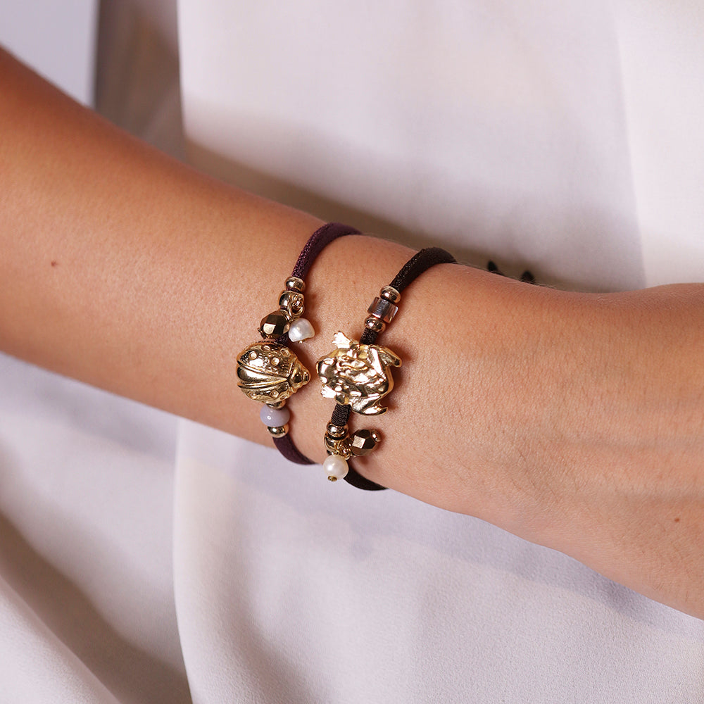 Pulsera de hilo marrón y morado con dijes dorados y cuentas blancas, estilo bohemio y elegante.