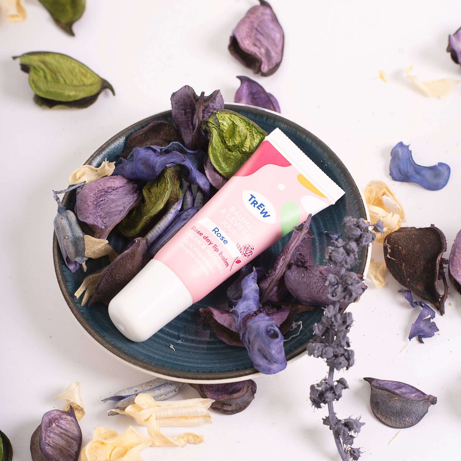 A pink and white lip balm tube resting on a small blue dish filled with dried purple and green flower petals.