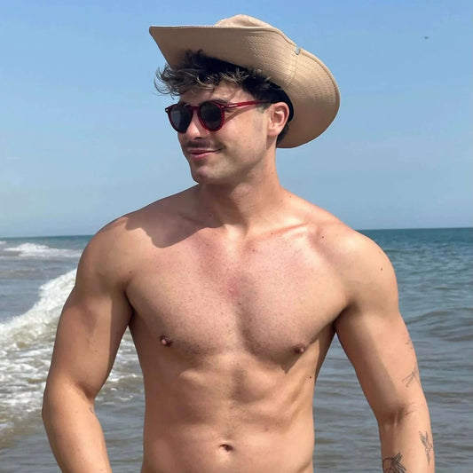 Hombre sin camiseta con sombrero en la playa usando gafas de sol redondas con montura y cristales en tono rojo intenso.