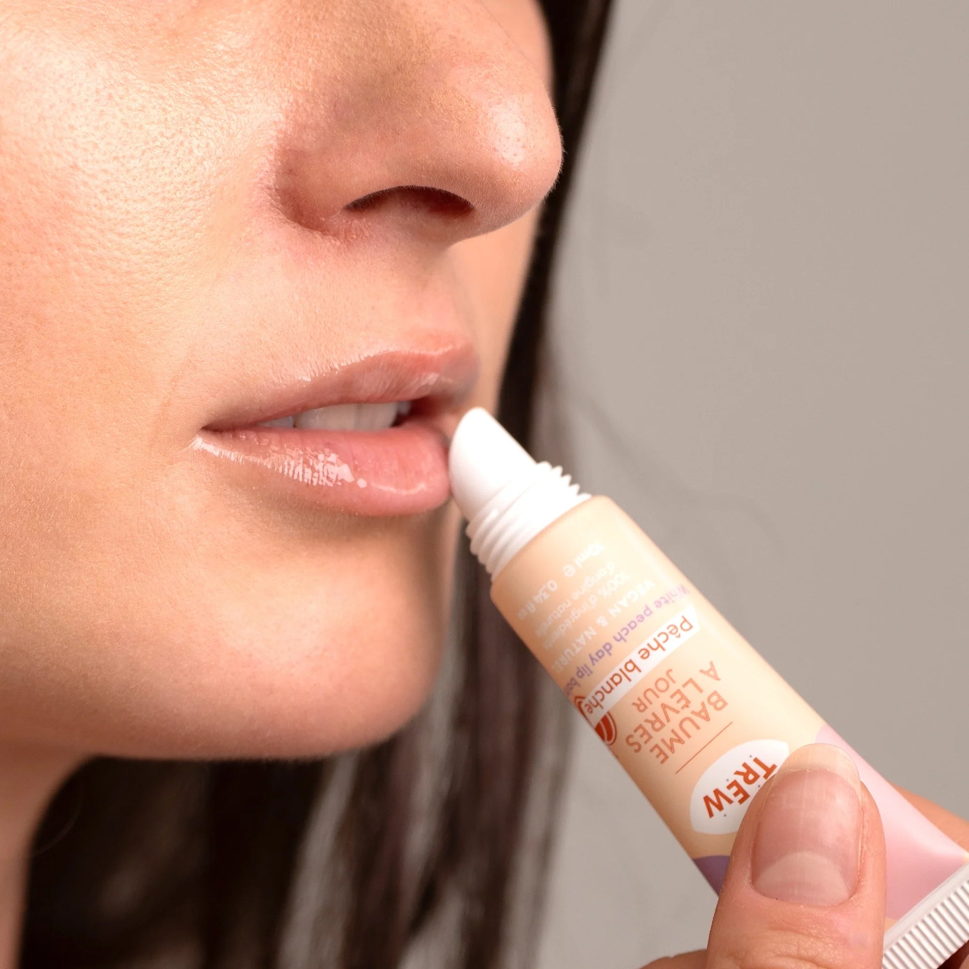 A person applies a peach-colored squeeze tube of TREW Baume à lèvres Pêche Blanche lip balm to their shiny, glossy lips.