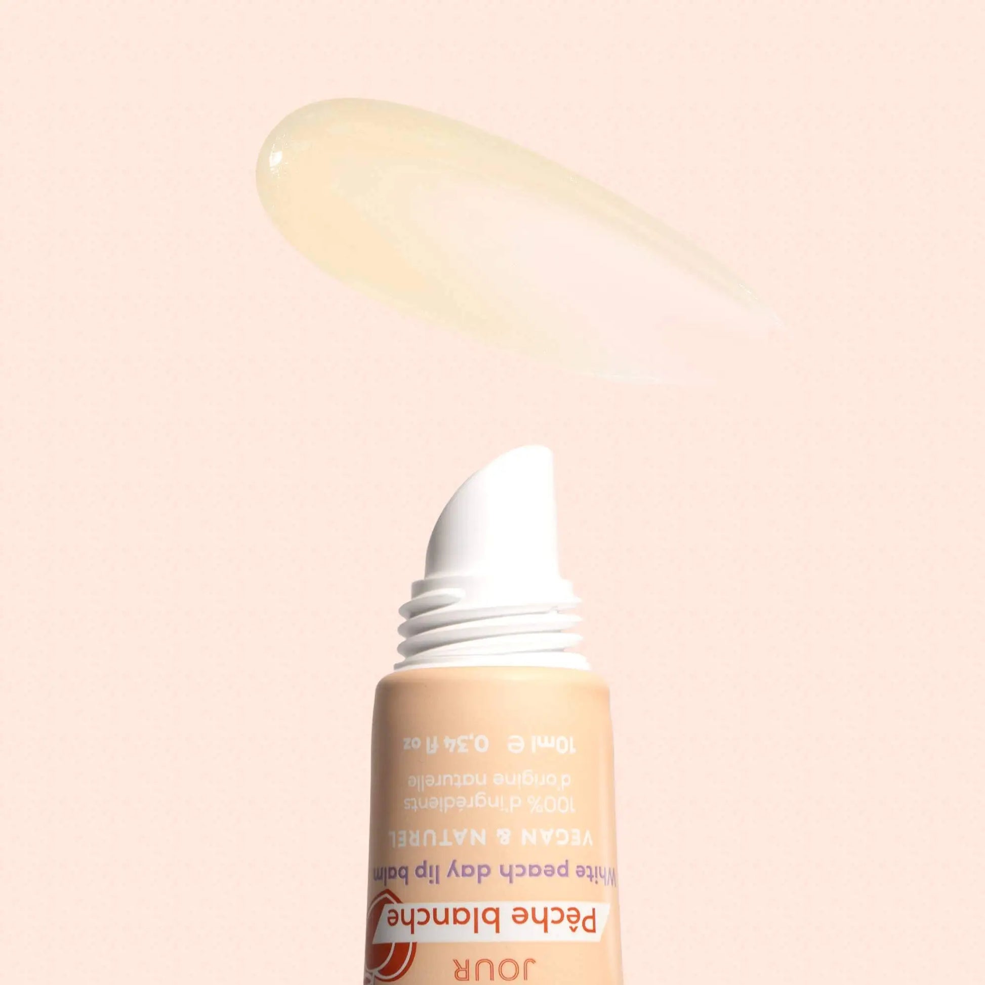 Squeeze tube of white peach lip balm with a smear of clear, glossy product above it on a soft peach background.